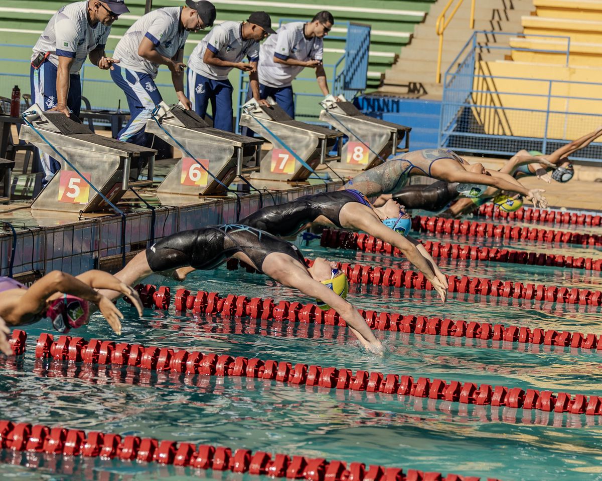 Piscina de competição durante largada de costas, com árbitros e arquibancada